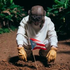 a man in white jacket and yellow gloves kneeling down holding an american flag on top of dirt