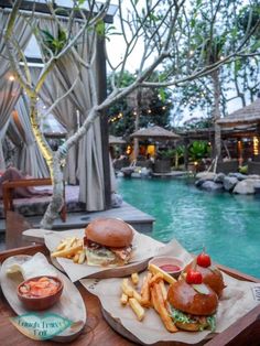 two burgers and fries are sitting on a table next to a pool at the resort