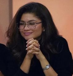 a woman wearing glasses sitting at a table with her hands folded in front of her face