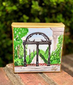 a small box with a painting on it sitting on top of a wooden table in front of some bushes