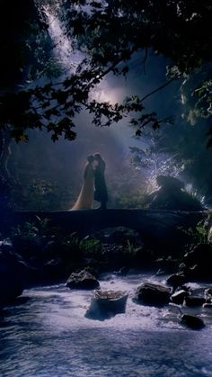 a man and woman standing on a bridge over a river in the woods at night