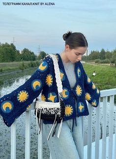 a woman standing on a bridge wearing a blue and yellow crochet sweater with sunflowers