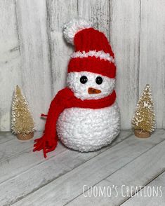 a stuffed snowman wearing a red hat and scarf next to small christmas trees on a white wooden surface