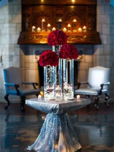 two tall vases filled with red roses on top of a table in front of a fireplace