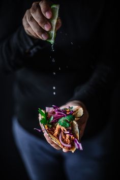 a person holding a sandwich with meat and vegetables being sprinkled on top of it