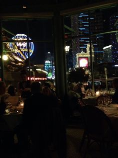 people are sitting at tables in a dimly lit restaurant with large windows overlooking the city