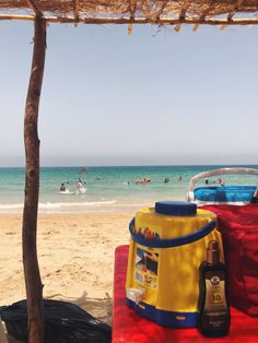 a yellow cooler sitting on top of a red blanket near the ocean with people swimming in the water