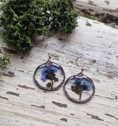 a pair of earrings with blue flowers in them sitting on a wooden table next to some plants