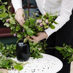 a woman arranging flowers in a vase on top of a white table with greenery