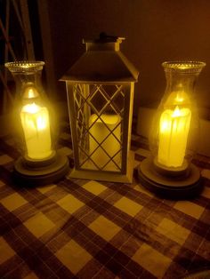 three lit candles sitting on top of a checkered tablecloth covered table with an old fashioned lantern