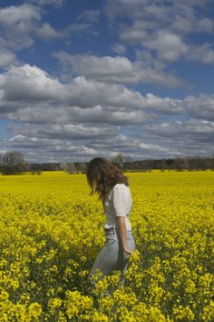 a woman walking through a field of yellow flowers under a blue sky with white clouds