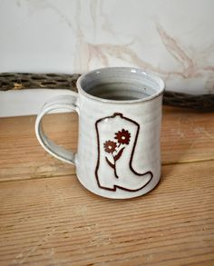 a white coffee mug with a cowboy boot and flower on it sitting on a wooden table