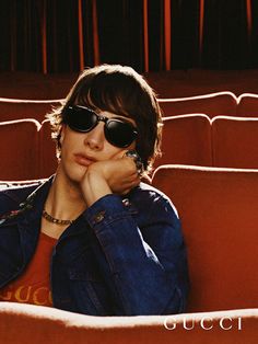 a man wearing sunglasses sitting in an auditorium with his hand on his chin and looking off to the side