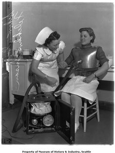 two women dressed in costumes sitting next to each other and one is using a hair dryer