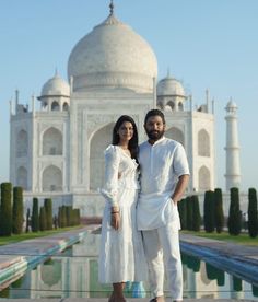 a man and woman standing in front of a white building with a reflection on the water