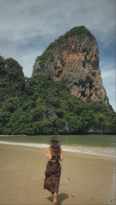 a woman walking on top of a sandy beach next to the ocean with a mountain in the background