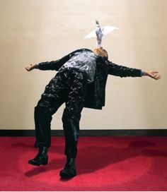 a man in black suit and white paper bird on his head standing on red carpet