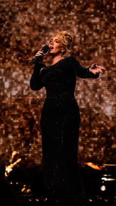 a woman in a black dress singing on stage with a microphone and lights behind her