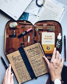 a person is writing on an open book with other items surrounding it and there are pens, notebooks, and papers all around them