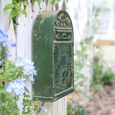 a green mailbox sitting on the side of a white fence next to blue flowers