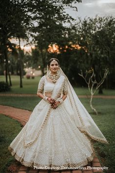 a woman in a white wedding gown standing on a brick path with trees and grass behind her