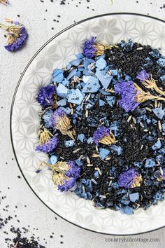 a white bowl filled with blue and purple flowers on top of a table next to dried herbs