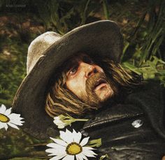 a man with long hair wearing a hat and holding daisies in his hand while laying on the ground