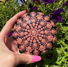 a hand holding an orange and black ball in front of some purple flower bushes