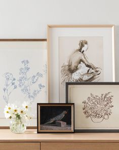 three framed pictures and two vases with flowers in them on a shelf next to artwork