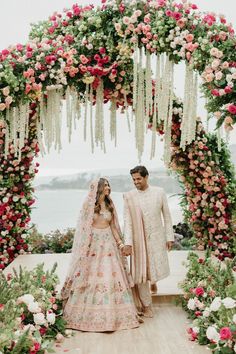 a bride and groom standing in front of a floral arch with flowers on it, holding hands