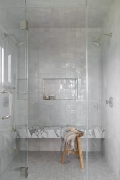 a walk in shower sitting next to a white tiled wall and floor with a wooden stool