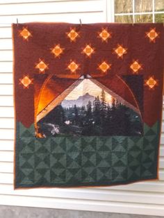 a quilt hanging on the side of a house with mountains and trees in the background