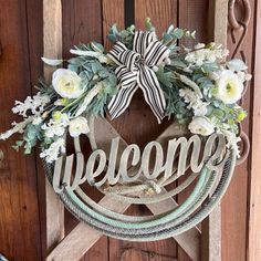 a wreath with the word welcome is hanging on a wooden door, decorated with white flowers and greenery