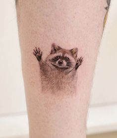 a small raccoon tattoo on the leg of a person's leg, with his hands in the air