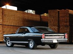 an old black car is parked in front of stacks of wood