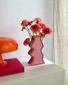 a pink vase with flowers sitting on top of a white table next to an orange lamp