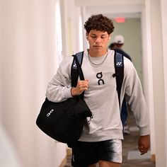 a young man walking down a hallway carrying a black bag and wearing a grey sweatshirt