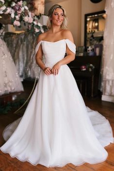 a woman wearing a white wedding dress standing in front of a floral arrangement and looking at the camera