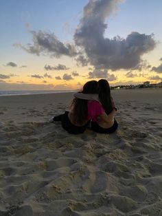 two women sitting in the sand at sunset with their arms around each other and looking into the distance