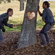 two men in the woods holding onto a tree with a heart shaped sign on it