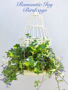 a white birdcage filled with green plants and purple flowers hanging from it's side