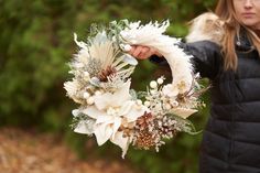 a woman holding a wreath with white flowers and feathers on it in front of trees