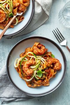 two plates filled with pasta and shrimp on top of a blue table cloth next to silverware