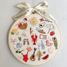 a white embroidered wall hanging with various items on it and a ribbon tied around the hoop