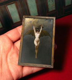 a hand holding a miniature painting of a nude woman with wings on her body and head