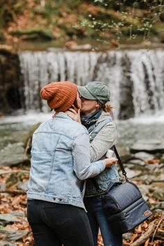 two women standing next to each other in front of a waterfall with leaves on the ground