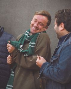 two men laughing while standing next to each other in front of a building with a green scarf around their neck