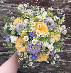 a bouquet of yellow and blue flowers is held by someone's hand in front of a stone wall