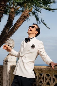 a man in a white tuxedo and bow tie standing next to a palm tree