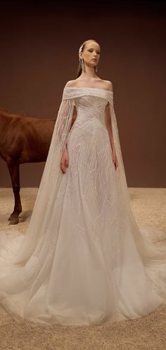 a woman standing next to a horse wearing a wedding dress with long sleeves and an off the shoulder cape